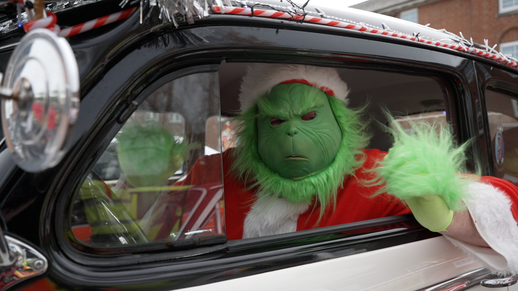 A man wearing a Santa's costume and a Grinch mask sat in a black classic car