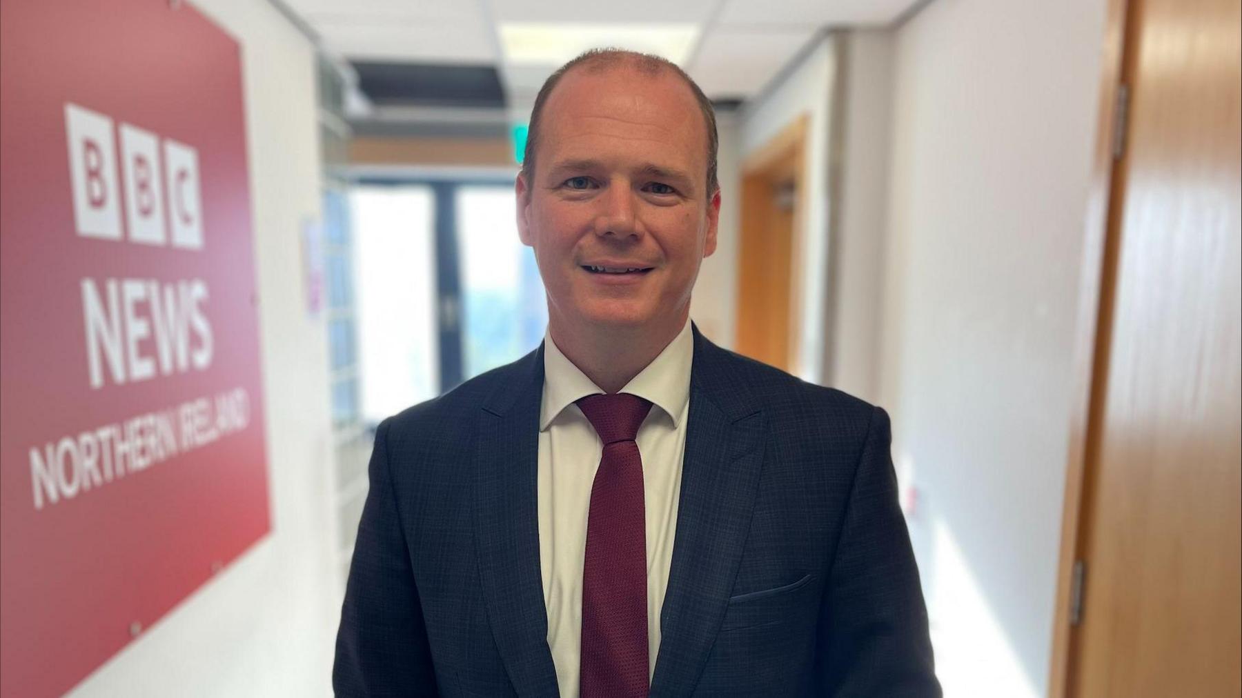 Sports Minister Gordon Lyons, wearing a navy suit, burgundy tie and white shirt. He is standing in a corridor with a red BBC News Northern Ireland sign on the wall to the left. 