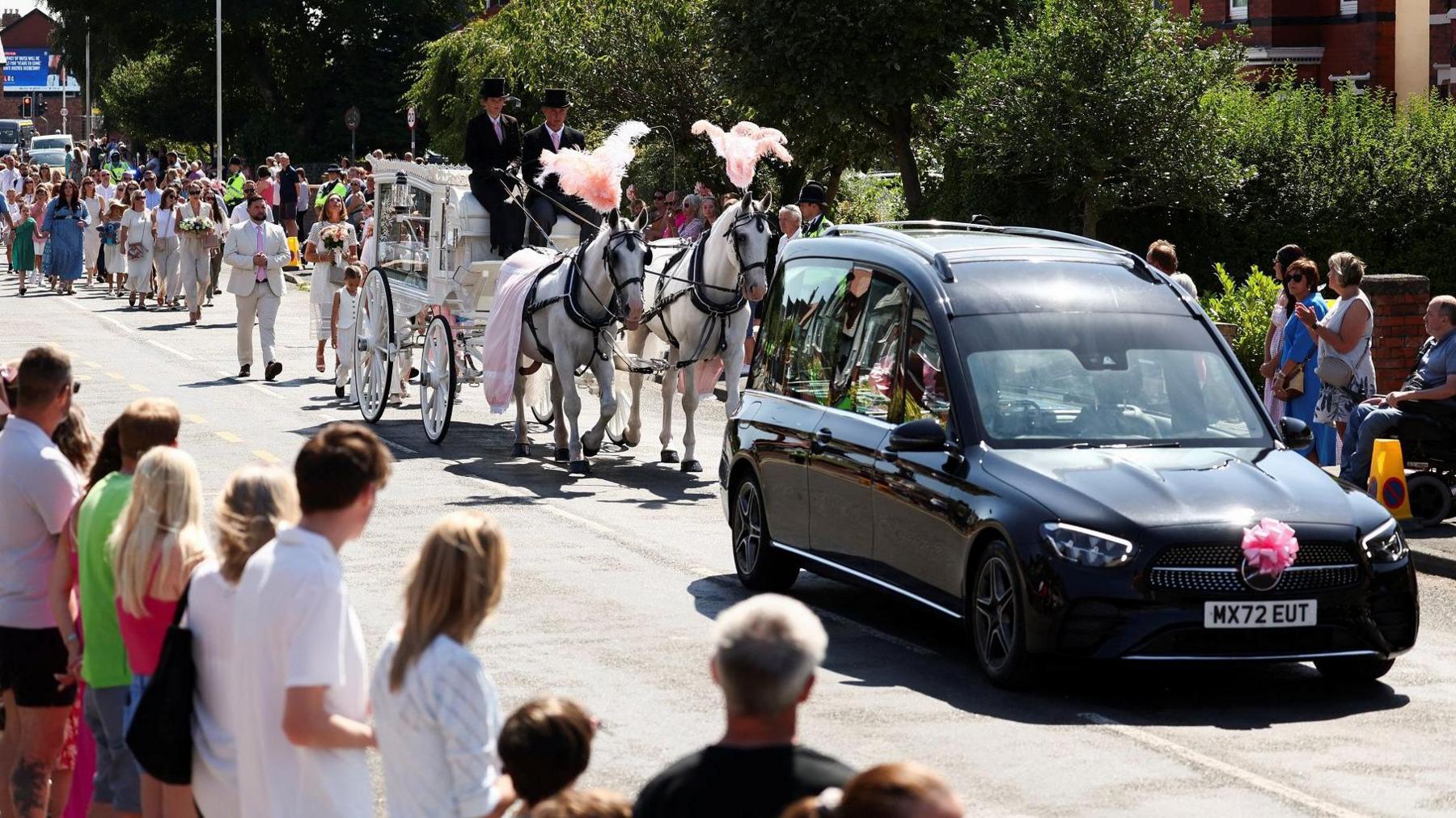 A funeral hearse and horse-drawn carriage leads the funeral procession as people watch from the sides of the road