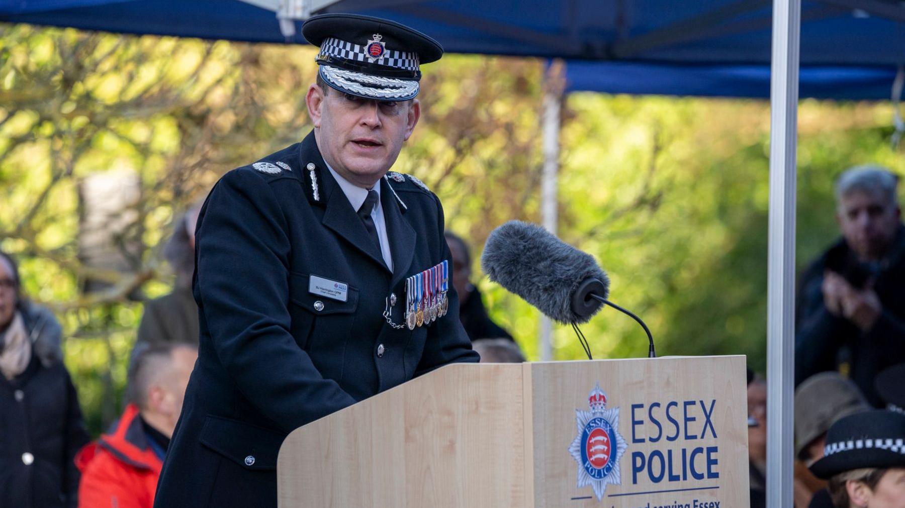 Ben-Julian Harrington wearing a black policing uniform and hat with a line of medals pinned to his chest. He is speaking behind a lectern which has a large microphone on it. People watch on behind him.