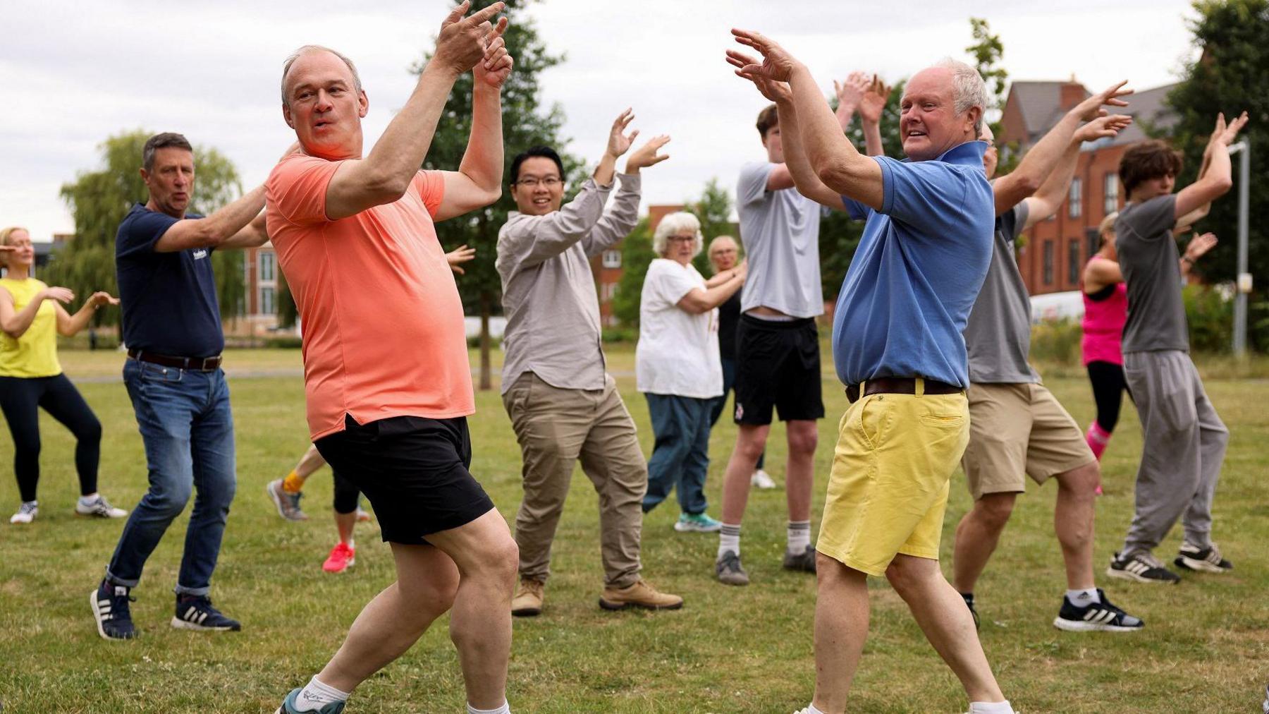 Clive Jones and Ed Davey wearing brightly coloured shorts and T-shirts, with their arms in the air taking part in a Zumba class with other participants around them