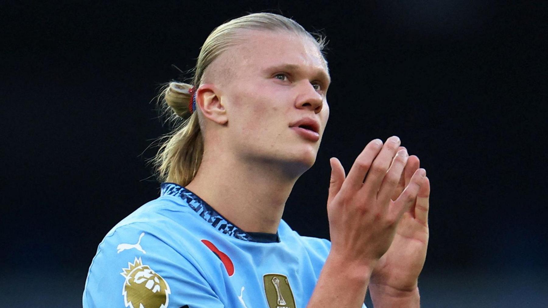 Erling Haaland holds his hands together as he looks to the heavens while playing for Manchester City. Wearing City's traditional sky blue shirt, Haaland sports his trademark ponytail.