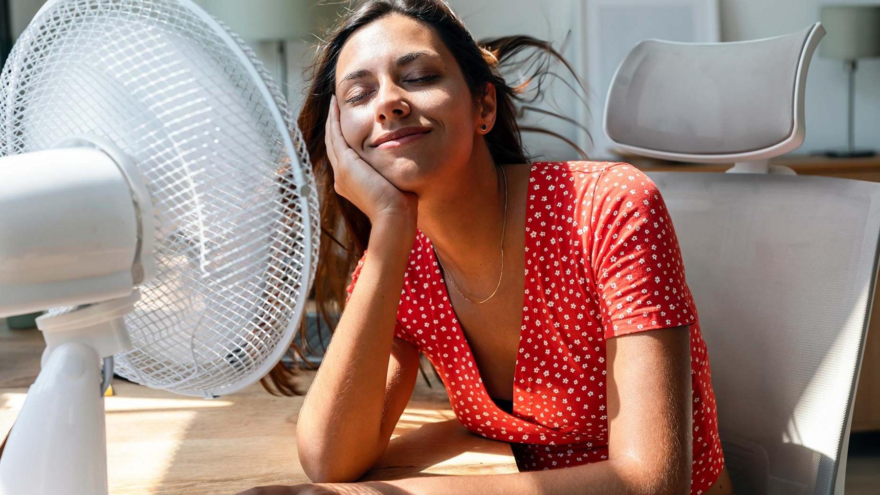 A woman keeps cool by a fan in her home wearing a red dress 
