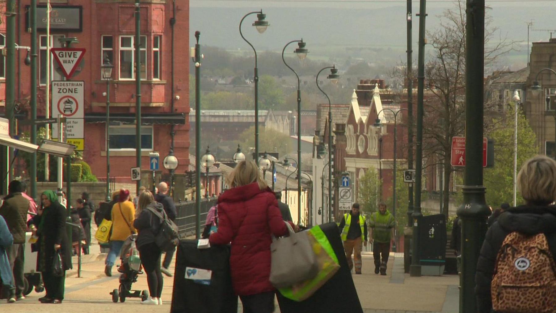 A woman in a red coat walks down a shopping street in Oldham clutching bags as other people mill around