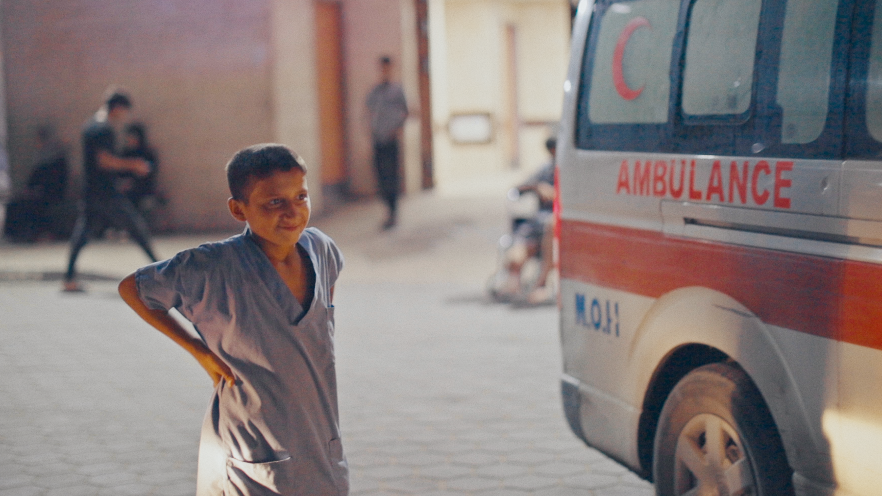 Zakaria wearing his blue scrubs as he stands next to a parked ambulance outside al-Aqsa hospital. He has his hands on his hips and is smiling as he looks towards the front of the vehicle.