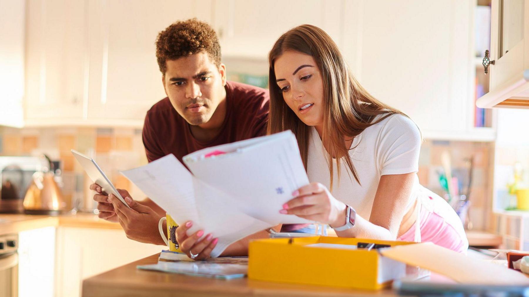 Couple leaning on kitchen worktop looking at bills with a yellow box folder on the counter in front of them