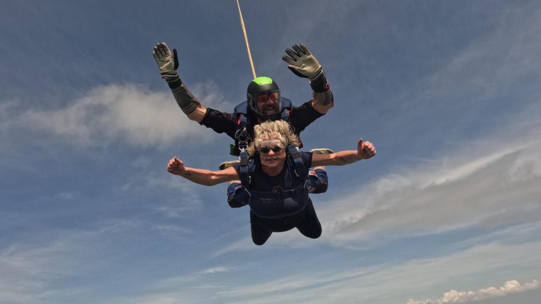 Lisa skydives in tandem with an instructor. She has her arms out to her sides and thumbs up.