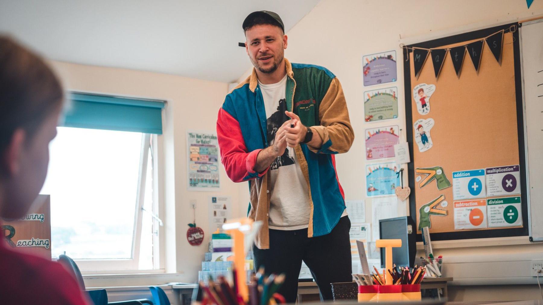 Connor in a class room setting. He is wearing a multi-coloured jacket and in the background you can see a class room board 