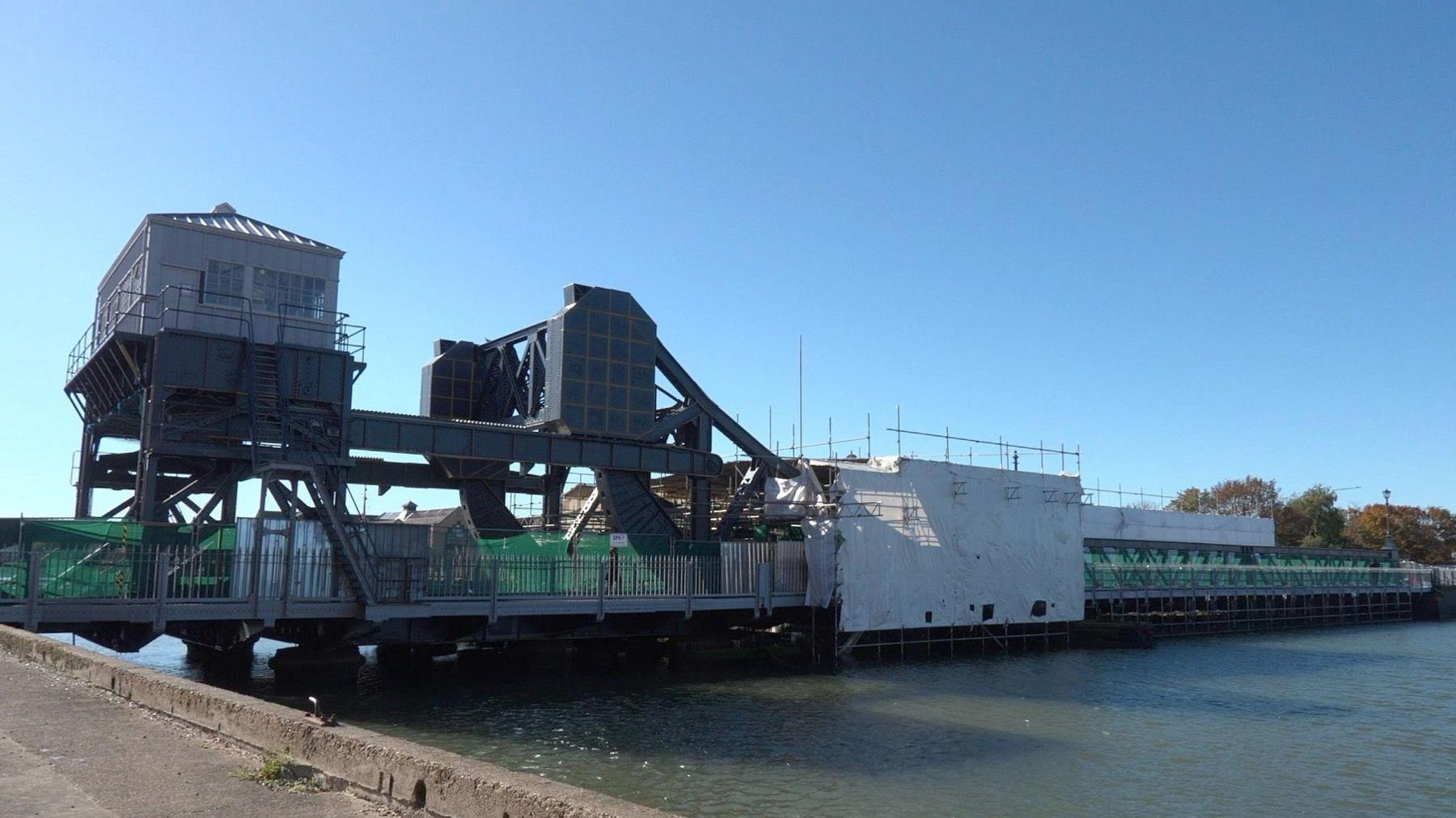 A 100-year-old bridge spans a wide river under a blue sky. It is seen sideways on from the bank and made of dark iron and steel. Much of the 150-metre (490-foot) span is short in height and sits just above the river, but on the left side an elaborate rolling lift structure rises to about 25 metres. Further to the left, another structure, also about 25 metres tall, houses a control cabin made of white wood and glass. Scaffolding and white canvas covers large parts of the bridge.