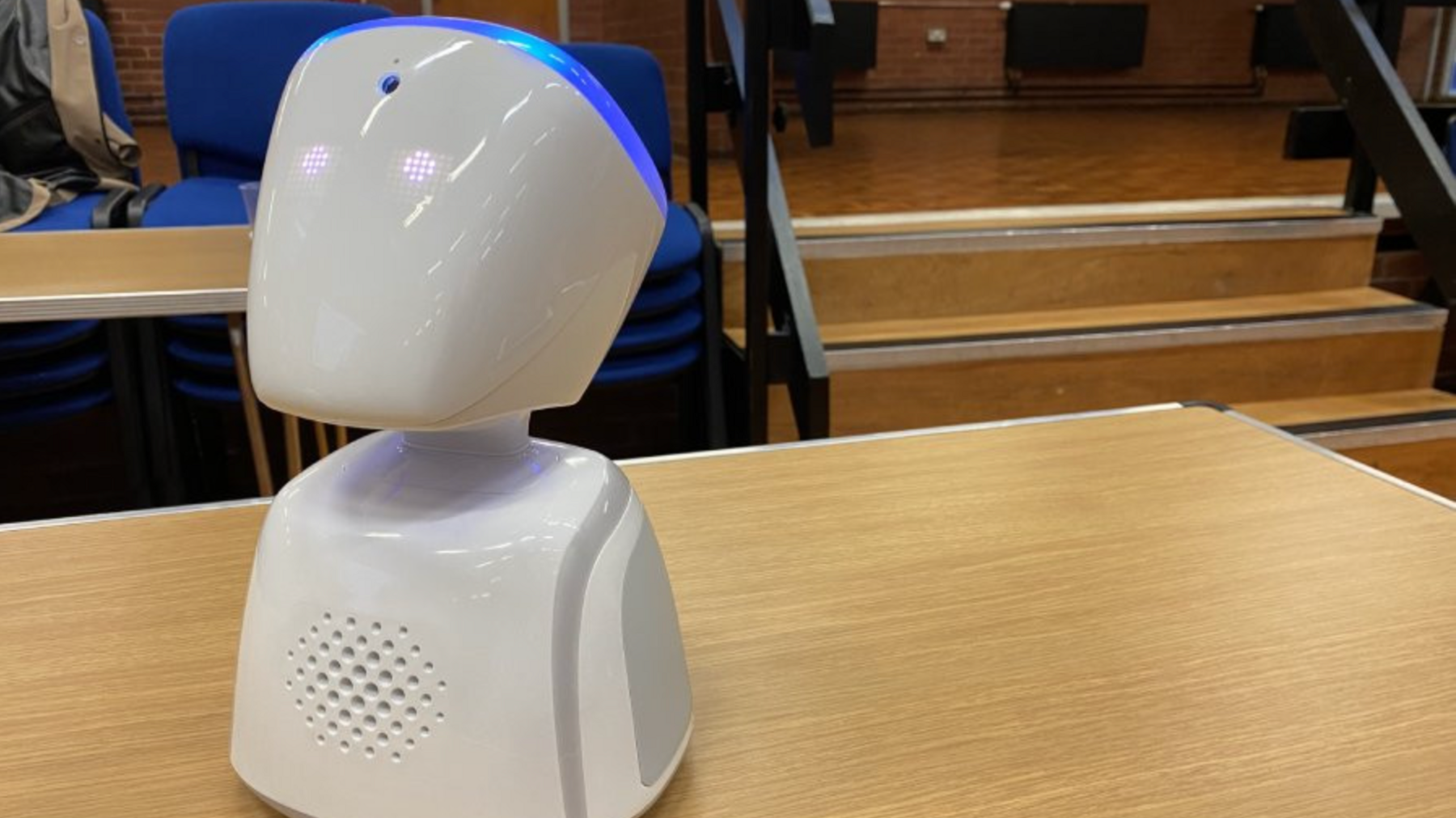 An image of a white robot sat on a wooden table in a school hall. It has glowing white eyes and a glowing blue strip on its head. 