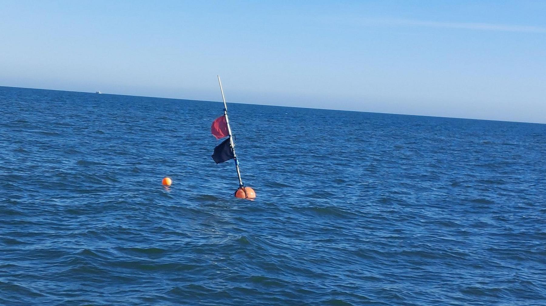 Photo of the open sea and a buoy in the middle with two flags - black and orange.