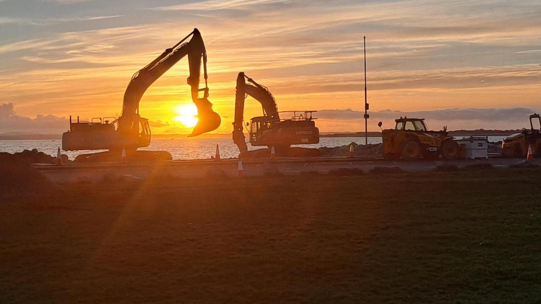 Construction vehicles are backlit by the setting sun as it heads towards the sea on the horizon. The photographer has carefully positioned the sun to appear to be in one of the excavators buckets.