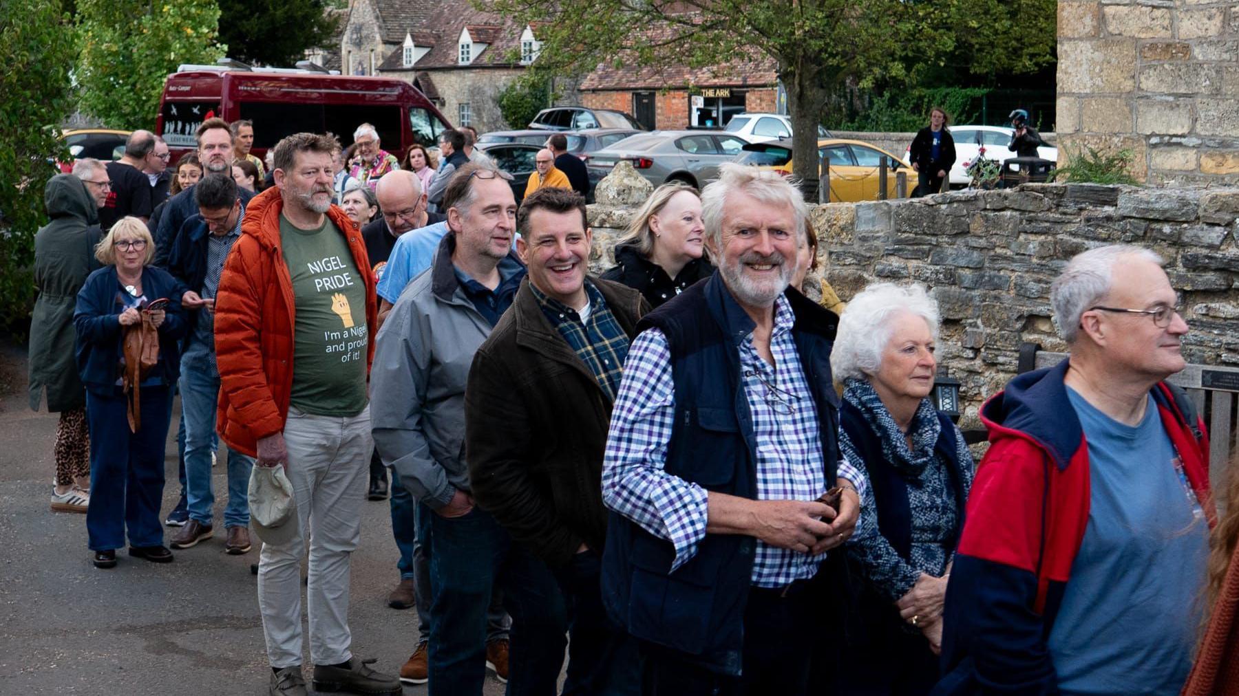 Men and women queue up at the pub for Nigel Night. The crowd is standing by a stone wall. There is a village with trees and cars in the background. 