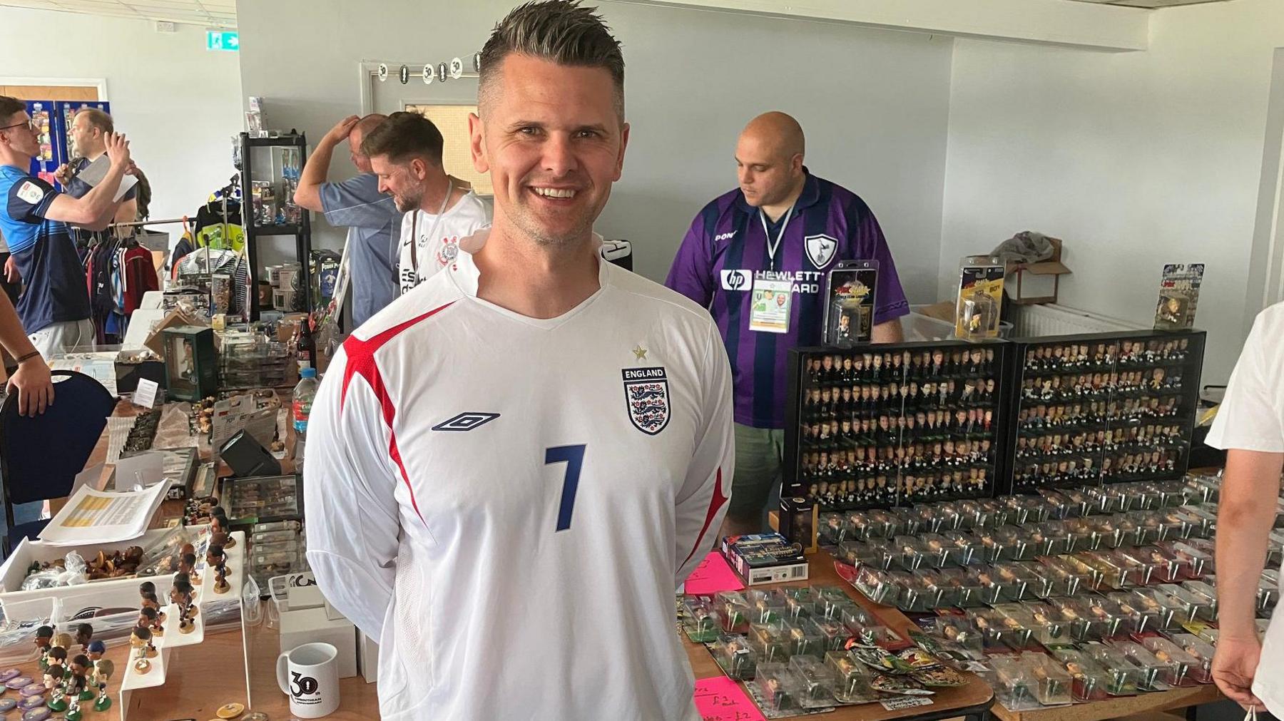 Adam Grocott smiles at the camera, wearing a white Umbro long sleeved England shirt with a red cross on the right shoulder and a blue number 7 in the middle. To his left is a table with hundreds of Corinthians models on, and more to his right. Other tables with memorabilia are behind and people manning the stand or milling about.