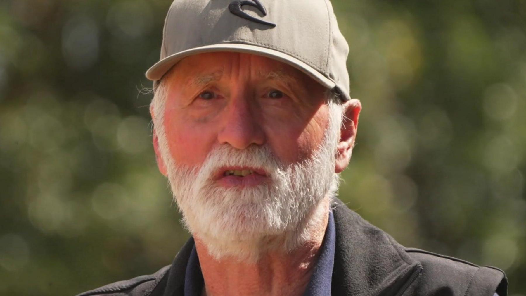 Nigel has a white beard and is wearing a grey cap and dark jacket.