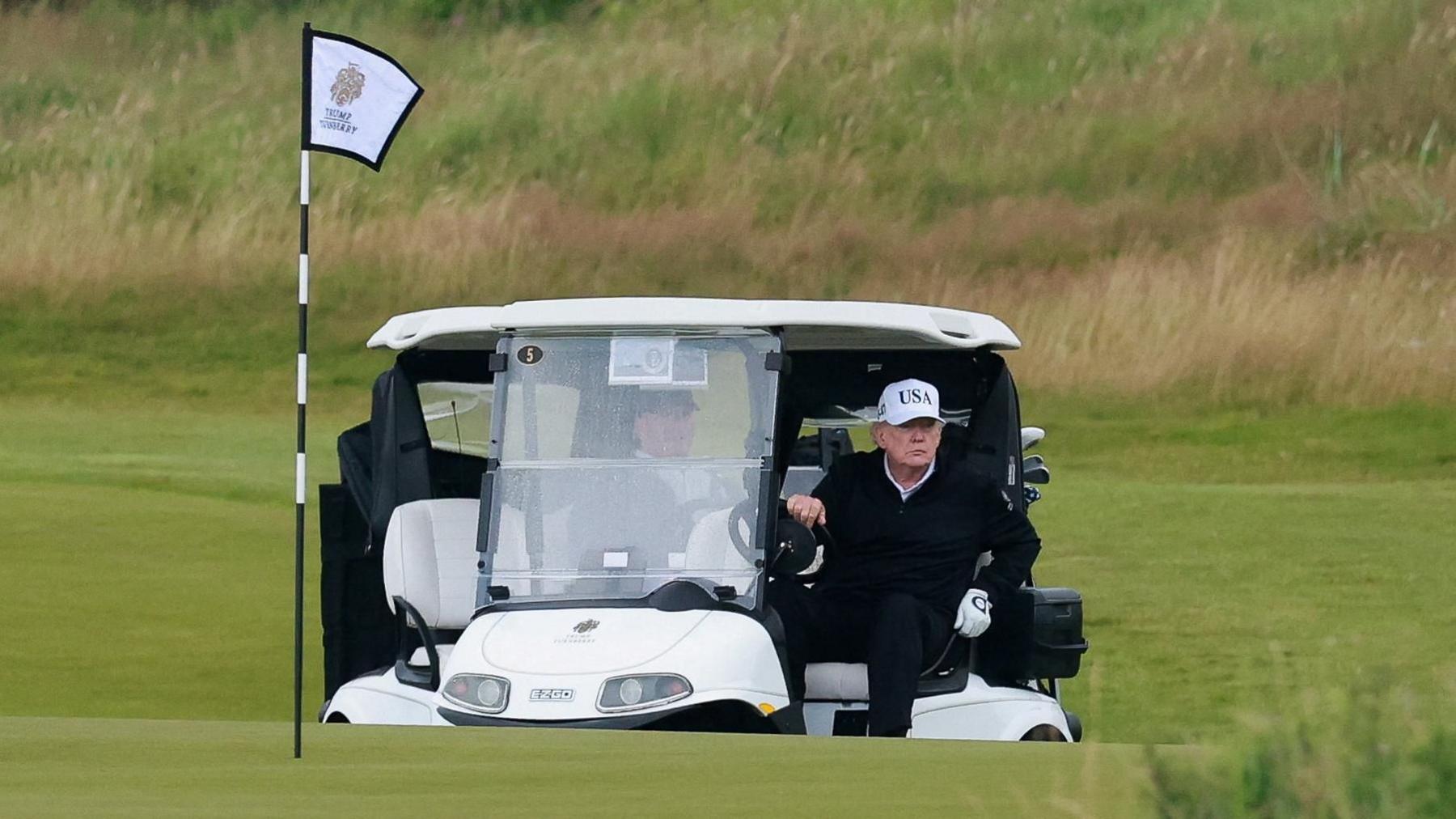 President Trump, wearing black clothes and a white baseball cap, gets out of a white golf buggy on the Turnberry course. There is a flag in the foreground.