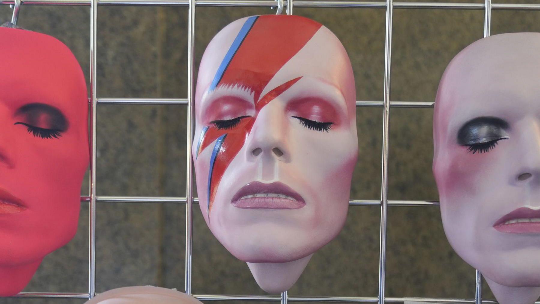 A mask of David Bowie's Ziggy Stardust character, with a red and blue lightning flash across it and eyes closed with long black eyelashes, is hung on a metal grid, alongside other masks of the singer in red and white tones