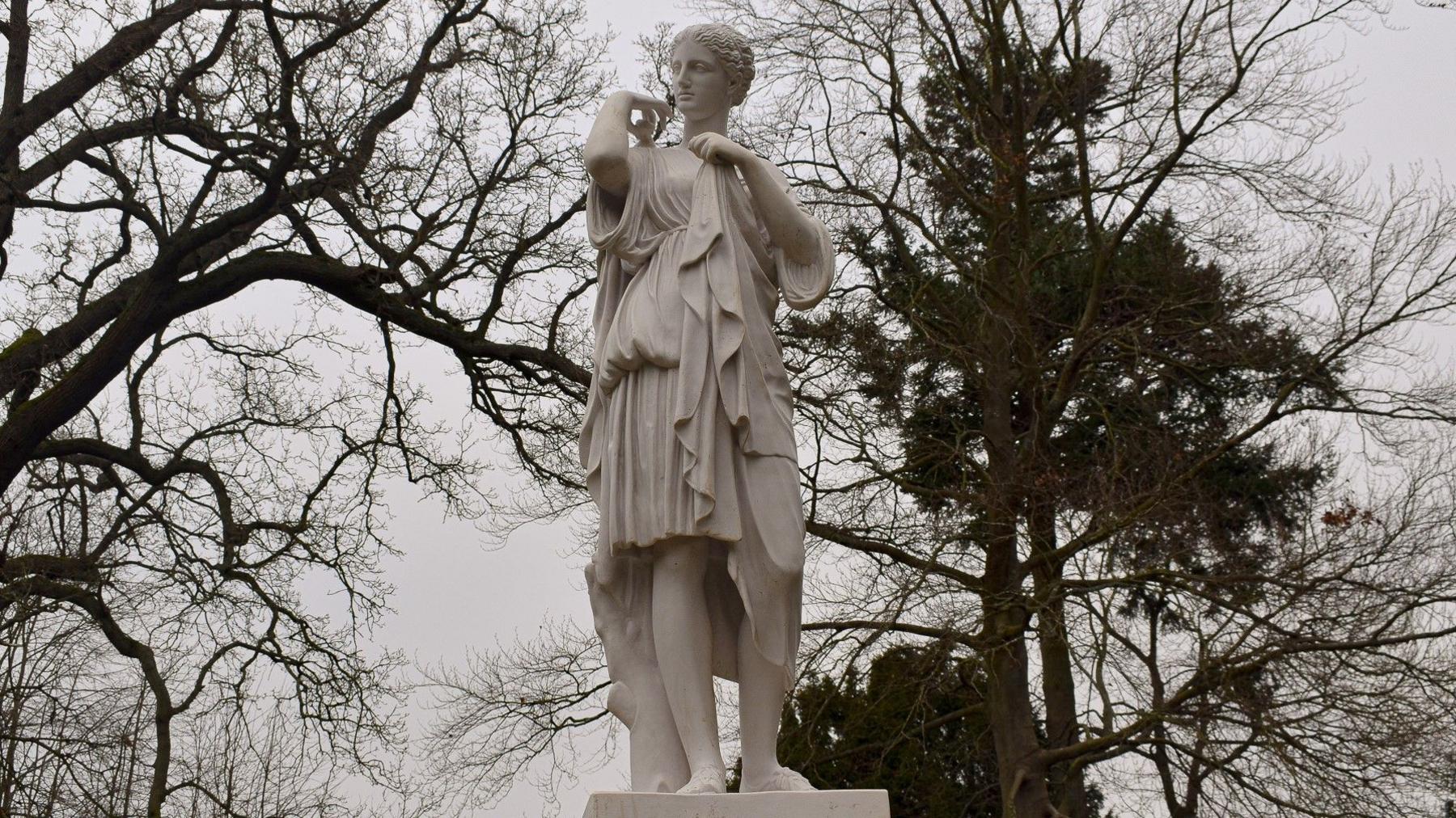 A statue of a woman clothed in draperies in a garden with trees in the background.