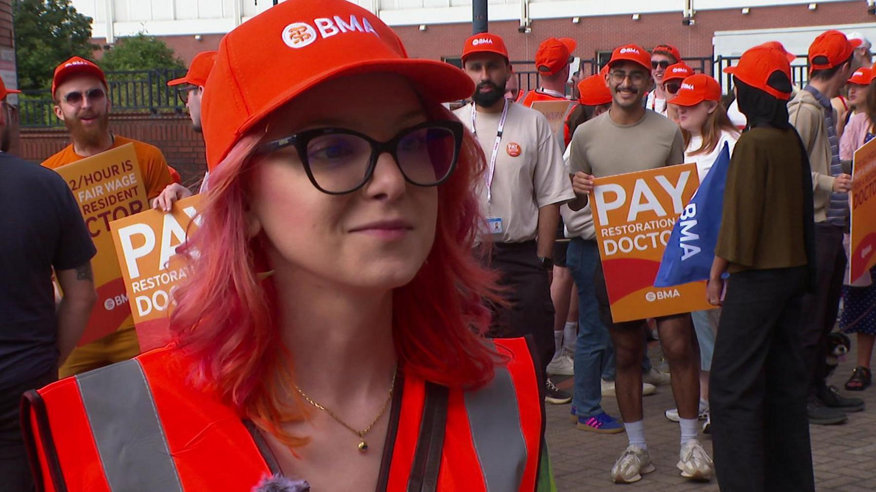 A woman wearing an orange cap with white lettering which reads BMA and a hi-vis jacket. She is wearing large black-rimmed spectacles and has red hair. Behind her are a number of people in similar hats holding placards.