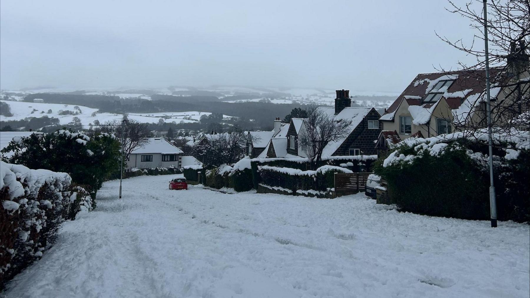 Heavy snow on a road with houses on each side and a red car in view. The moors are in the background, covered in snow.