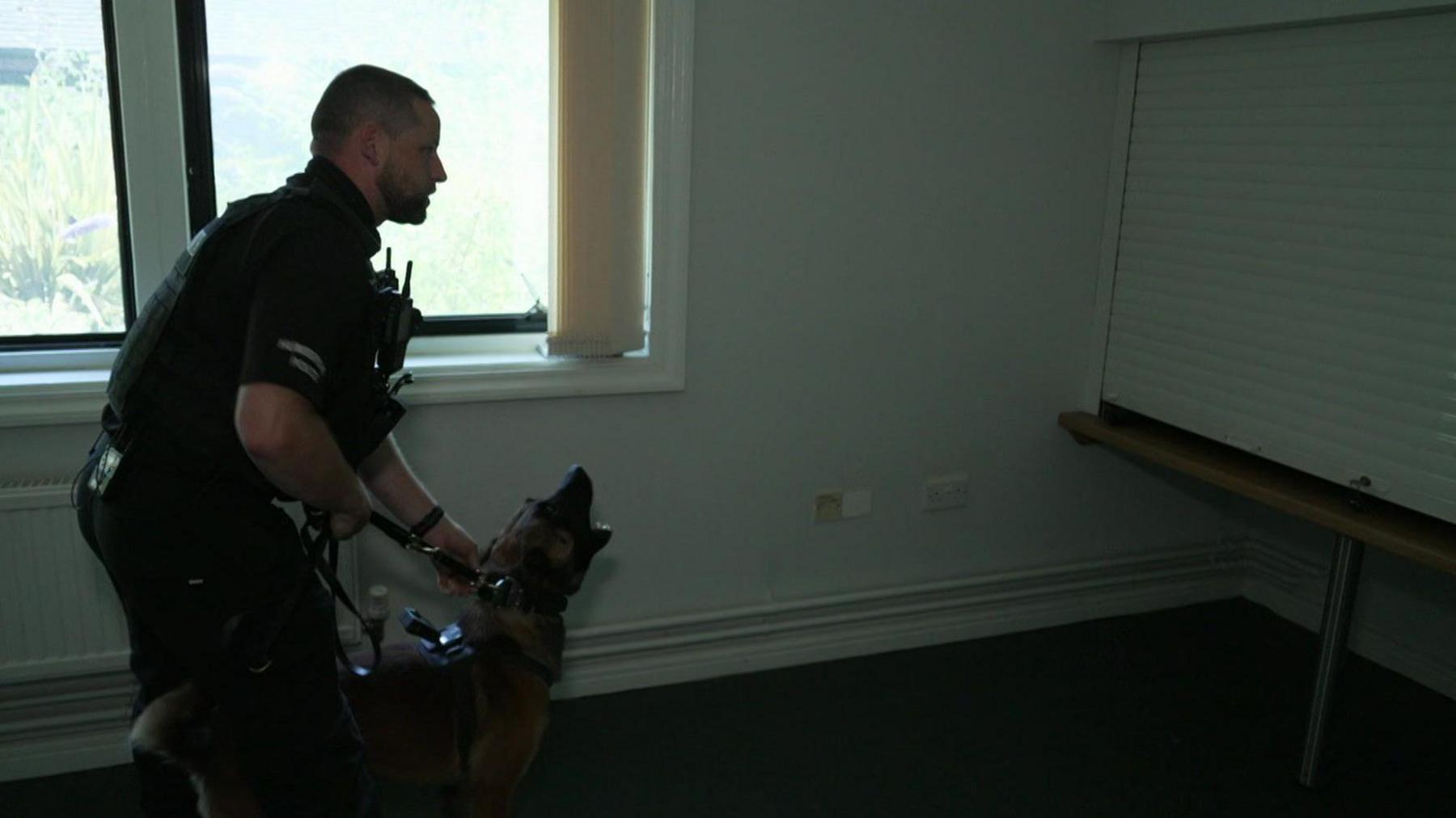 A police officer holds back his dog, a Belgian Malinois. Journalist Kevin Shoesmith is out of sight, hiding inside a raised cupboard 