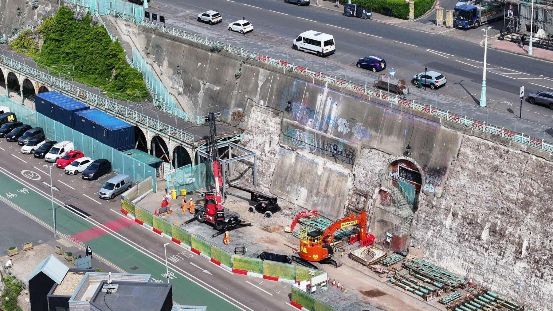 A large section of concrete wall as seen from the air with blue railings and some of the blue archway pieces laid to one side next to bright orange construction equipment on Brighton promenade