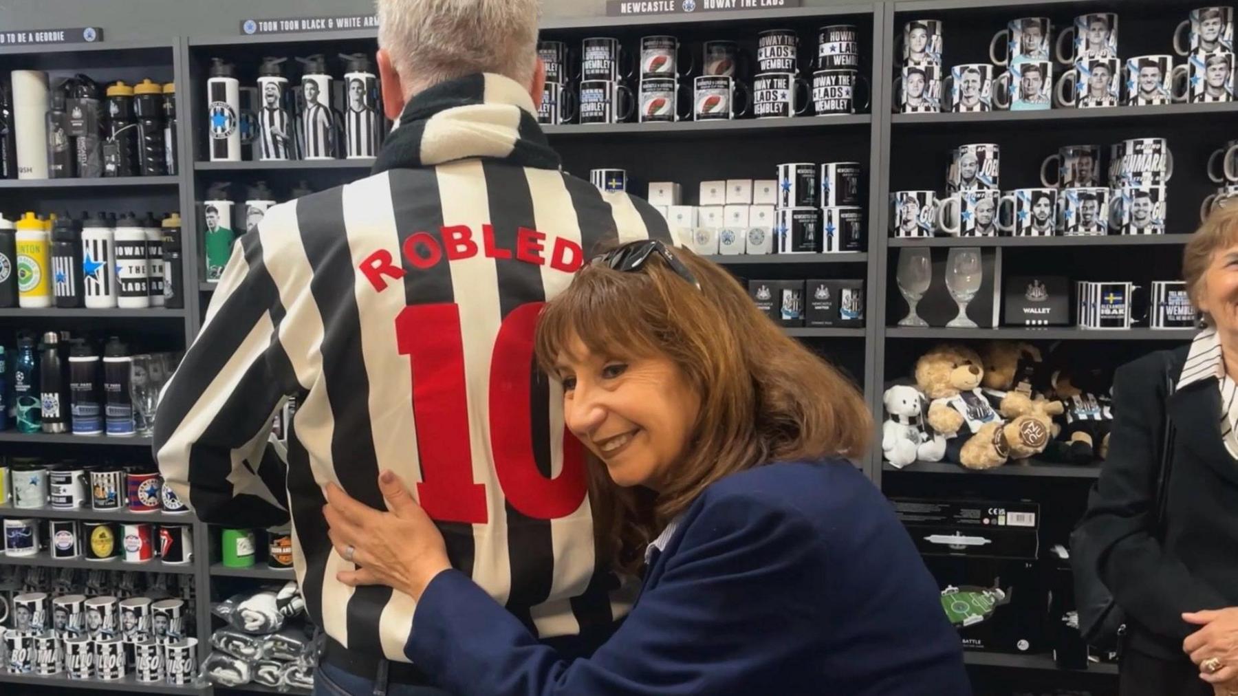 Elizabeth Robledo hugging a man who is facing away from her so only his back is visible. He is wearing a black-and-white football shirt with "Robledo, 10" on the back. Behind them are shelves stacked with football memorabilia.