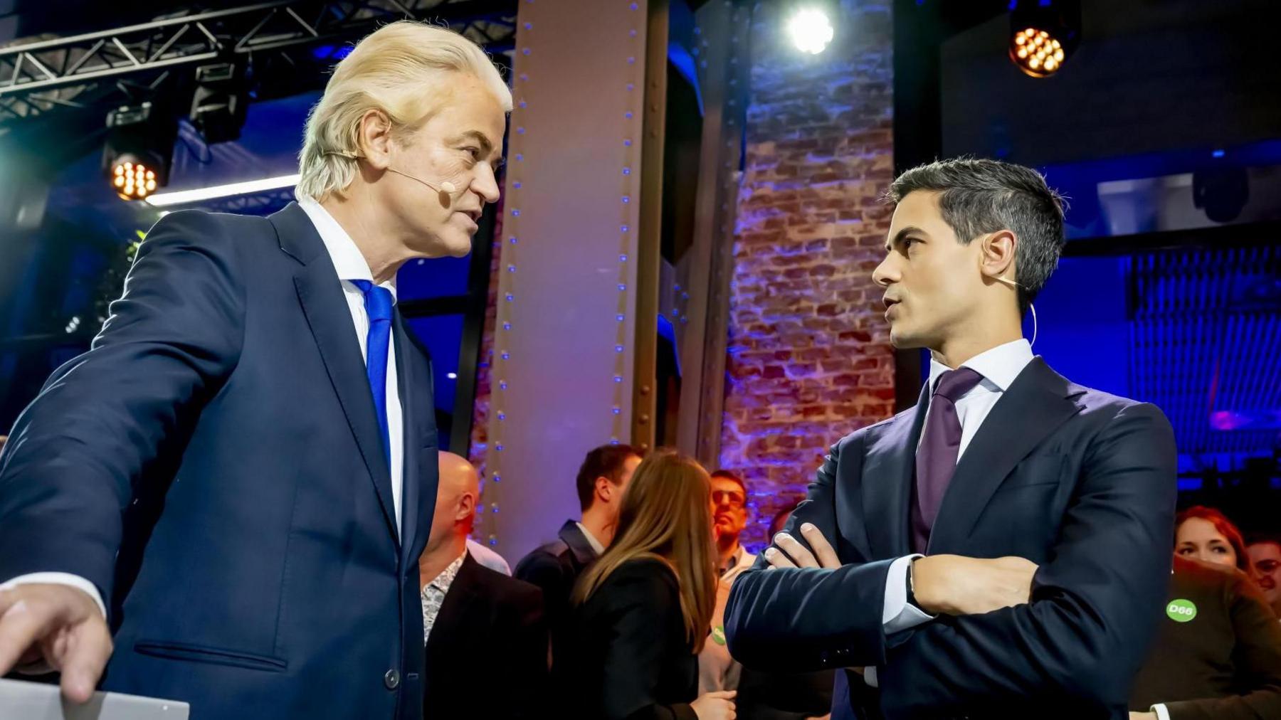 Geert Wilders and Rob Jetten, both wearing suits, speak to each other at a debate event.