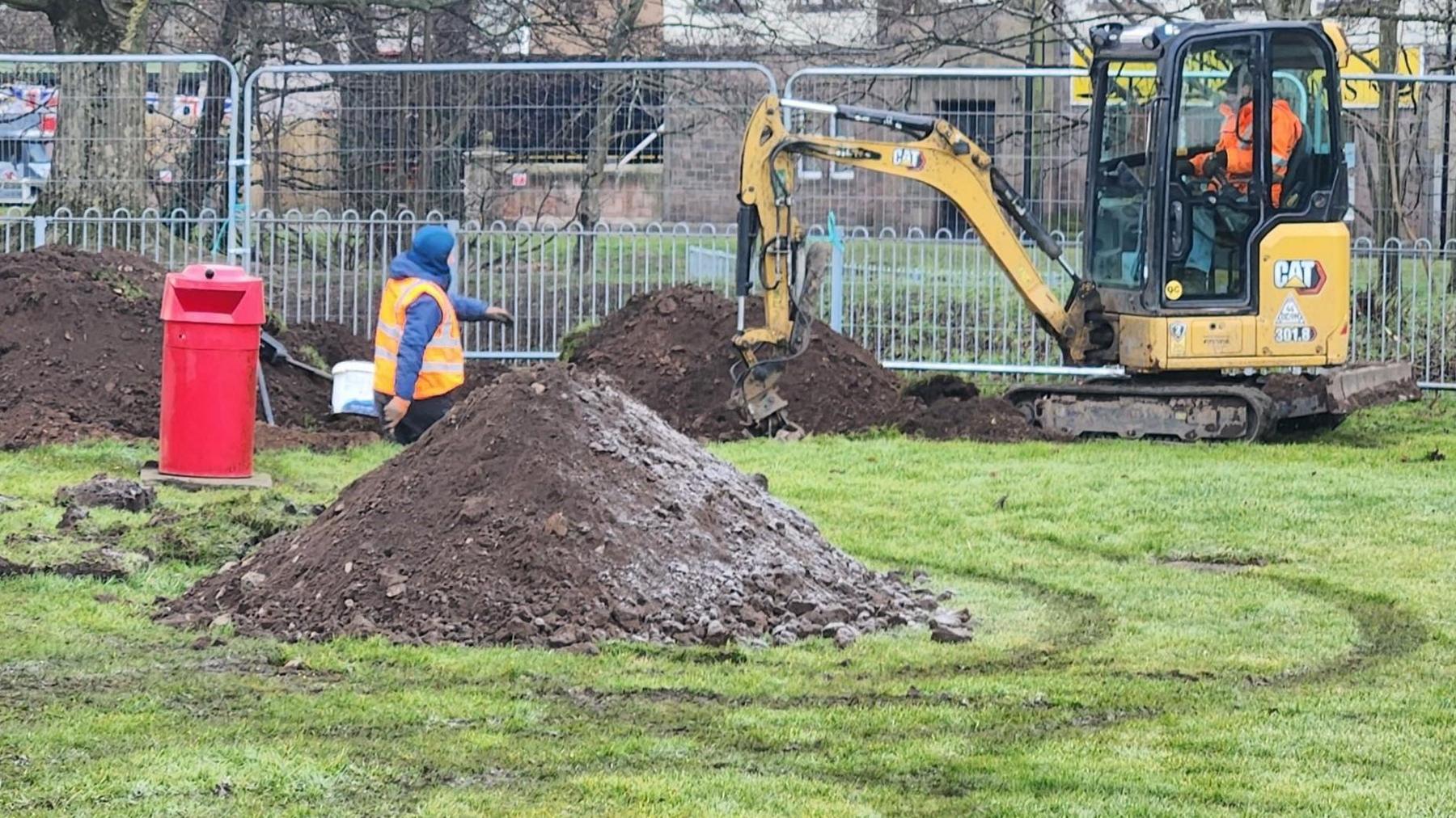 A digger is at work in a play park with big railings behind 