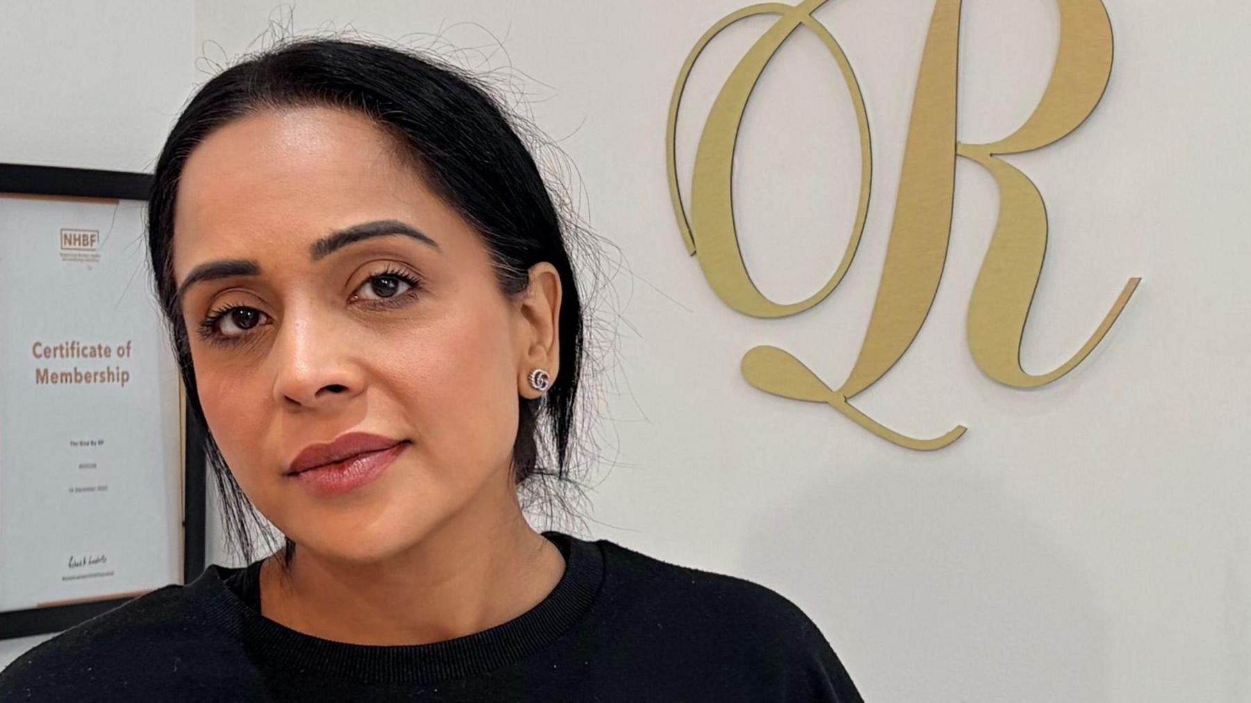 A woman in a black sweater stands behind a counter and looks at the camera. A gold telephone, two cards and a white plant are on the desk. Two gold letters - 'r' and 'p' are hung up on the wall alongside a certificate.