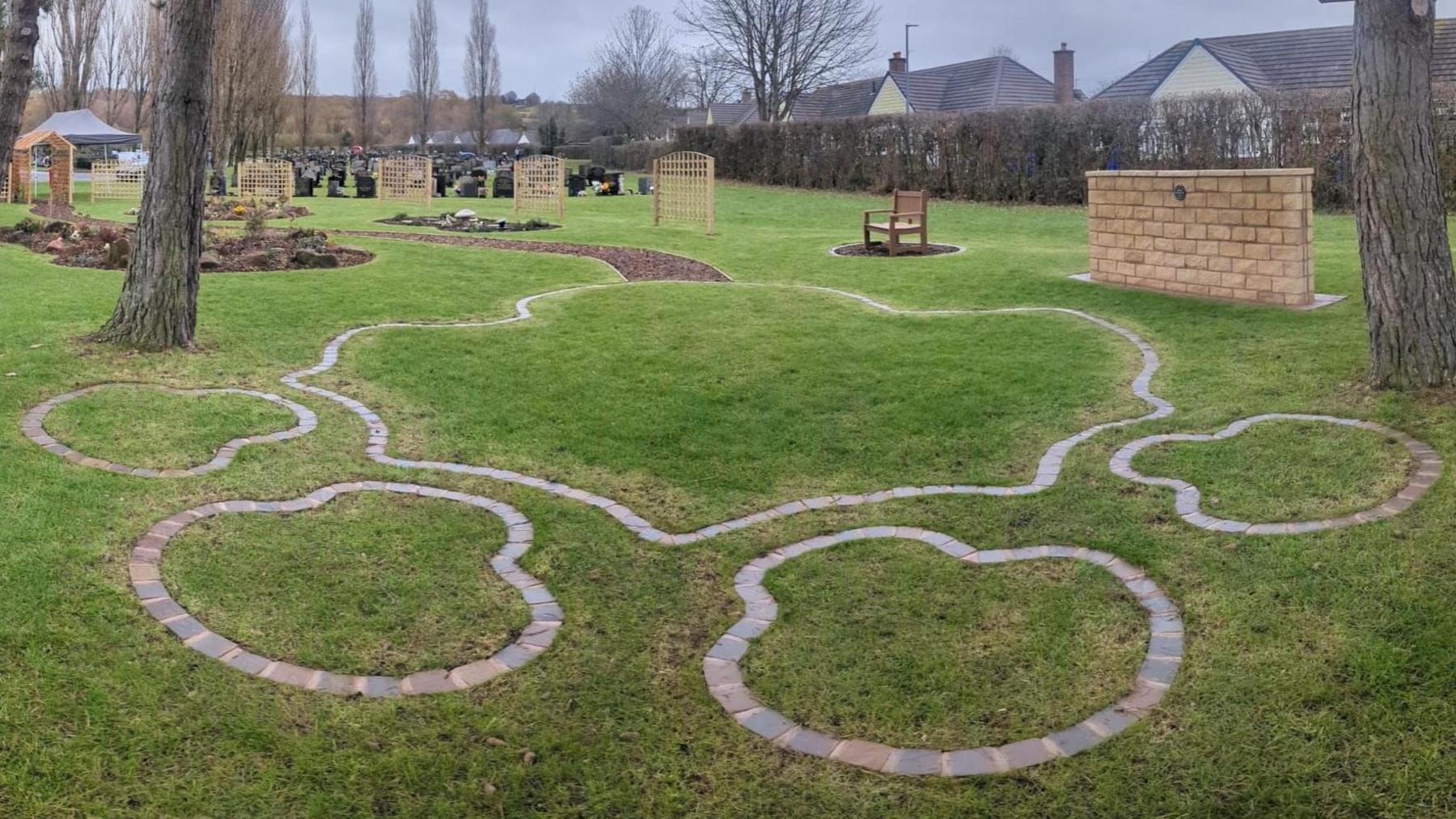 A garden with bricks laid to create the outline of an animal paw. There is a brick wall and a woodchip path as well as wooden trellises.