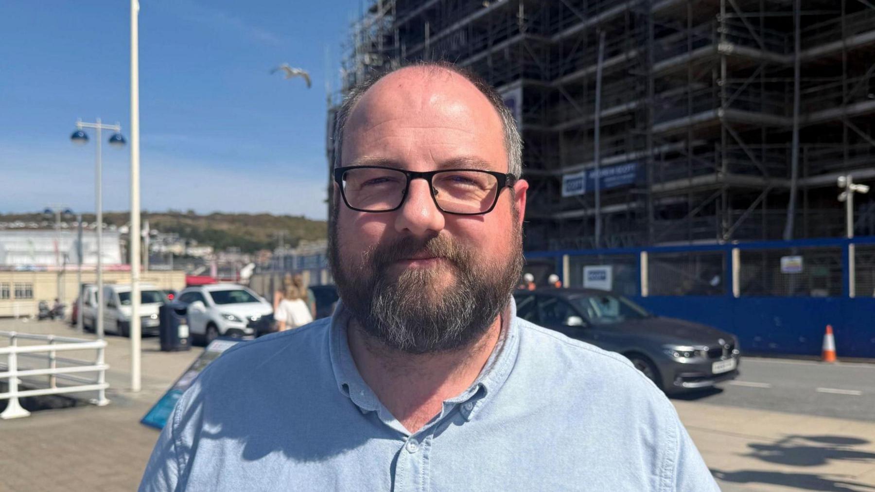 Emlyn Jones, in a blue button down Oxford shirt looks ahead into the camera, standing in front of the scaffolding that covers part of the Old College building.