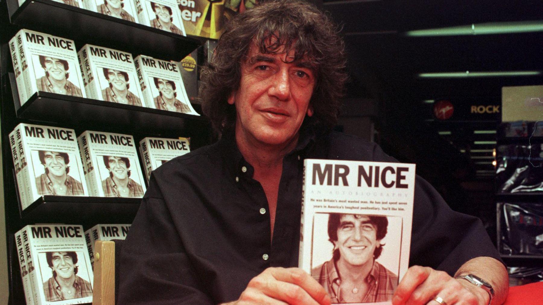 Howard Marks holding a copy of "Mr Nice". He has almost chin-length dark curly hair and is wearing a black shirt. 