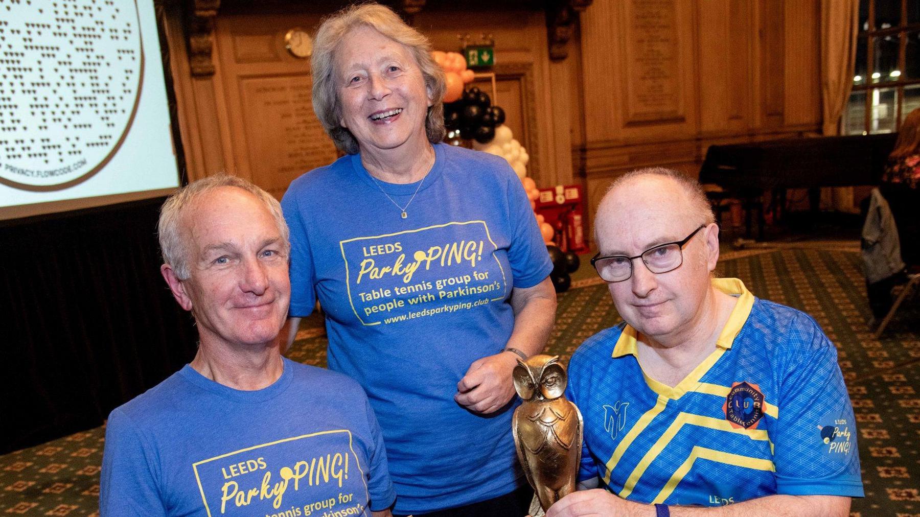 Three people are seated and standing at a table in a formal indoor venue with ornate wood-panelled walls and patterned carpet. They are wearing blue shirts, two of which display the text “Leeds ParkyPING! Table tennis group for people with Parkinson’s.” Rob is holding a bronze owl-shaped trophy.
