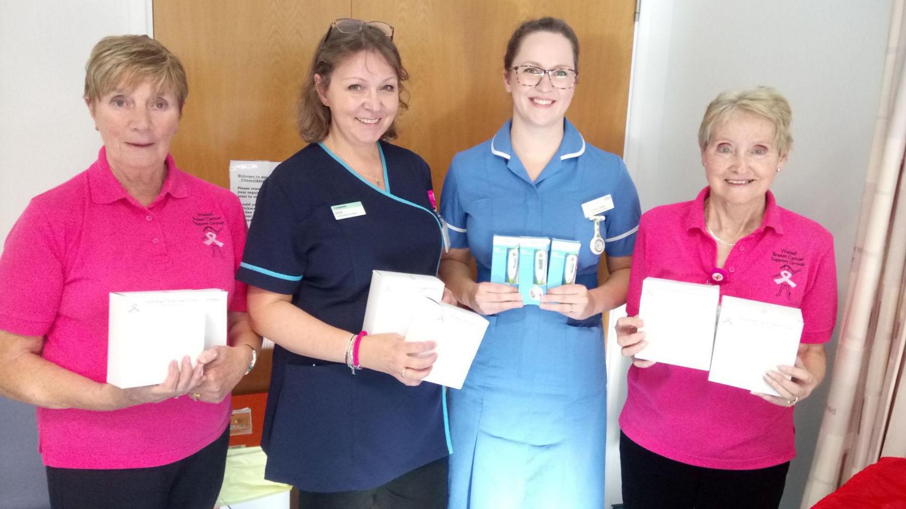 Four women stand in a row smiling as they hold up white boxes. The women on each end wear bright pink t-shirts with a pink breast cancer ribbon printed on the breast. The two in the centre are wearing blue nurse uniforms. they are standing in a hospital ward