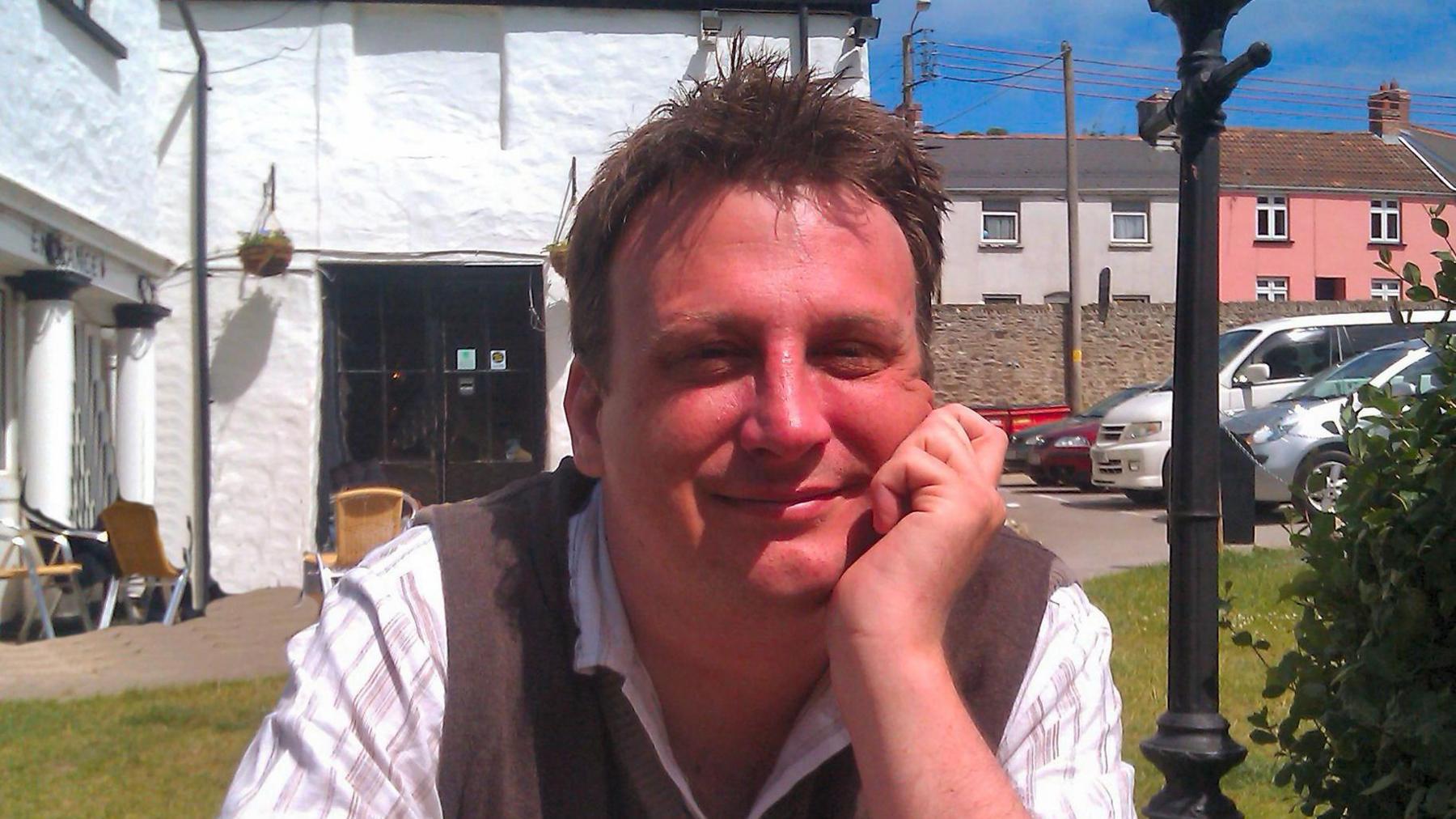 Nick Howard, 51, has brown spikey hair, he is wearing a brown tank top over a white shirt with brown pin stripes in. He is sitting down at a bench with his chin resting on his left cupped hand, behind him are cars parked and a white building with a grass lawn in the foreground.
