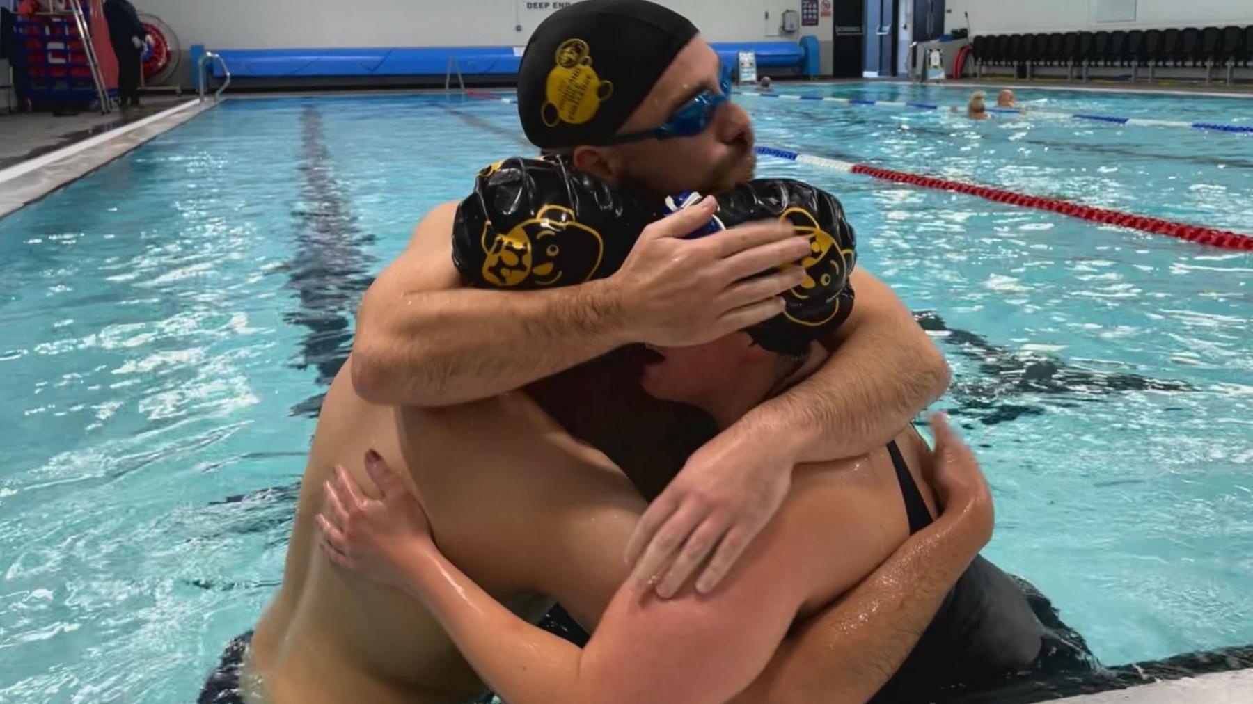 Two men and one woman wearing swimming caps stand and embrace in a swimming pool.