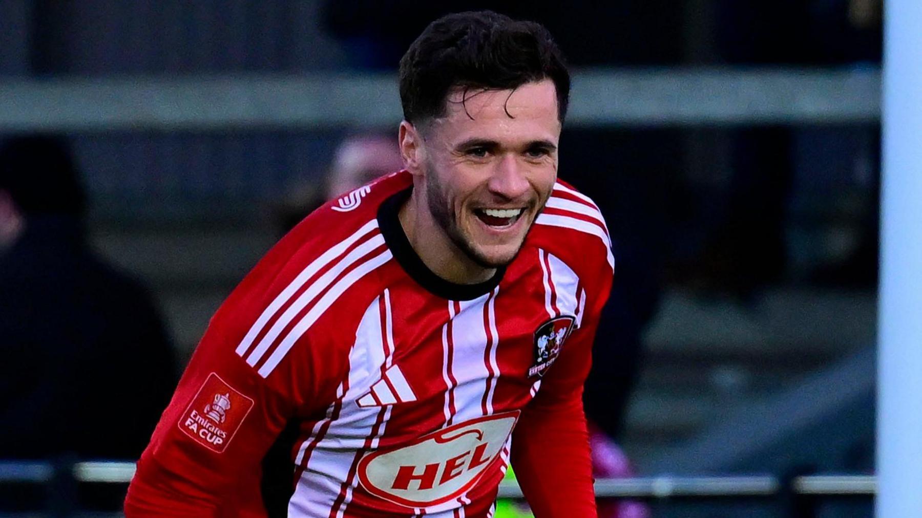 Jack Aitchison with short black hair and a neat beard is smiling. He is wearing th Exeter City home strip with red and white stripes.