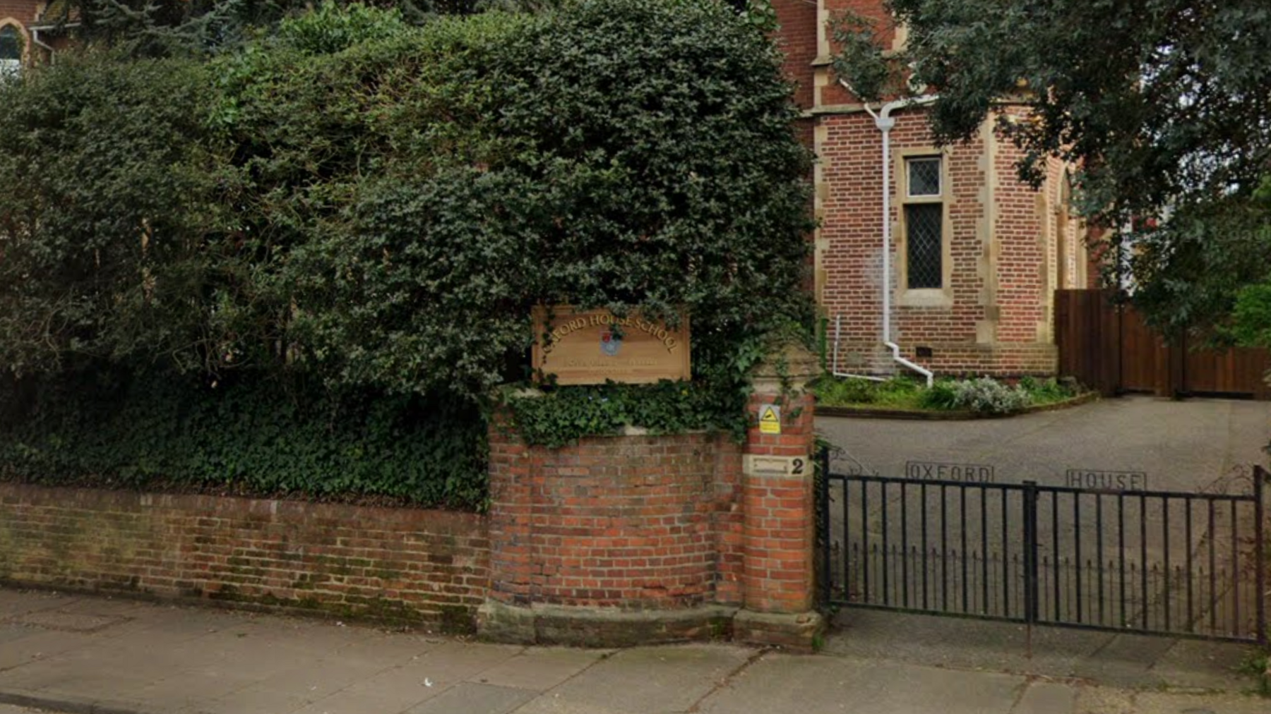 A brick wall and foliage behind it with a wooden sign with the words "Oxford House School" in front of a school building. To the right of the sign is a black metal gate with the words "Oxford House" on top.