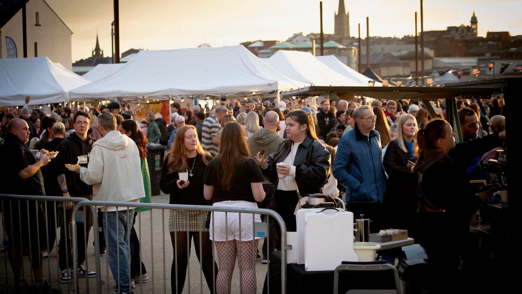A crowd of people are gathered. They are eating, drinking and talking. There are white marquee tents in the background. 