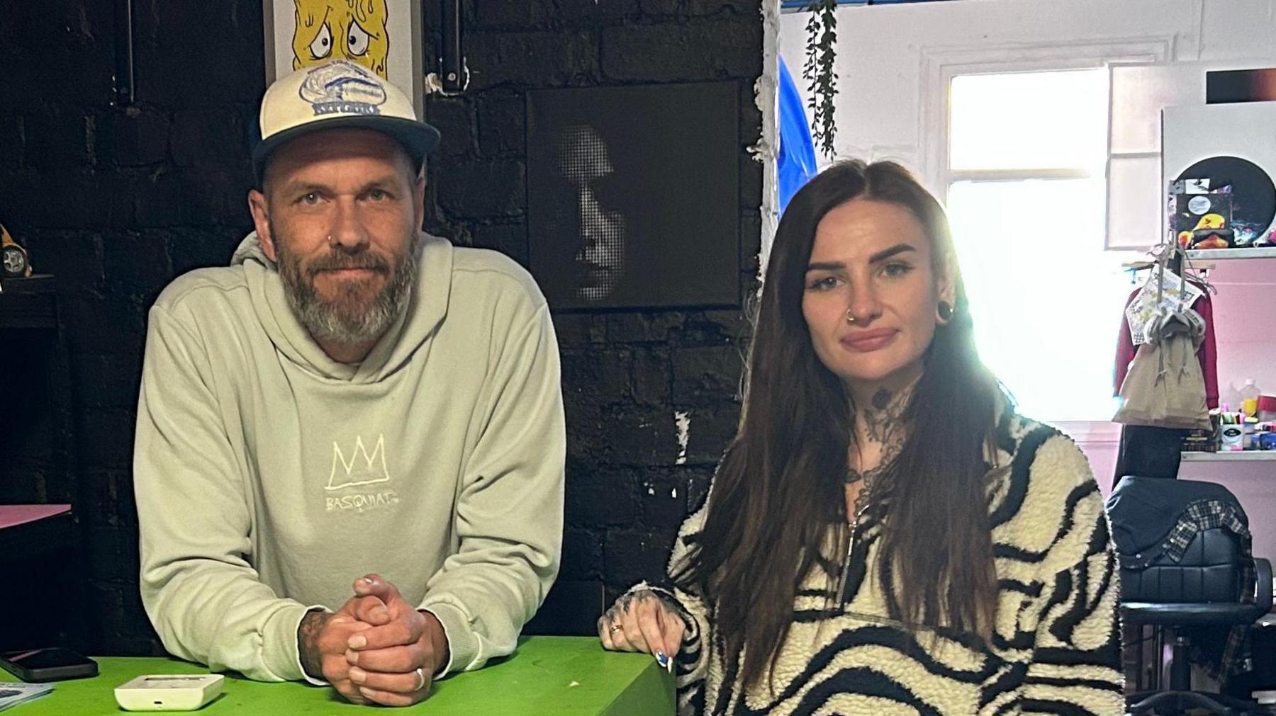 Mark James and Daisy Farrar-Hayton, co-owners of Irregular Art Hub stand side-by-side. Mark is in a light coloured hoodie and a baseball cap and learning over a counter. Daisy is in a black and white zebra print patterned fleece. She has a nose ring, tattoos on her neck and long brown hair. They are in a shop.