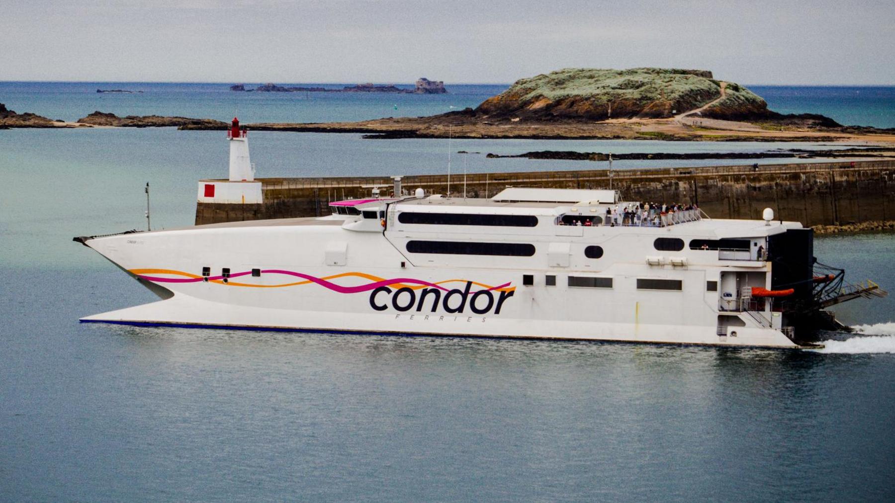 A ferry - small, white vessel with Condor written on the side with two pink and orange wavy lines - near a dock with an island in the background