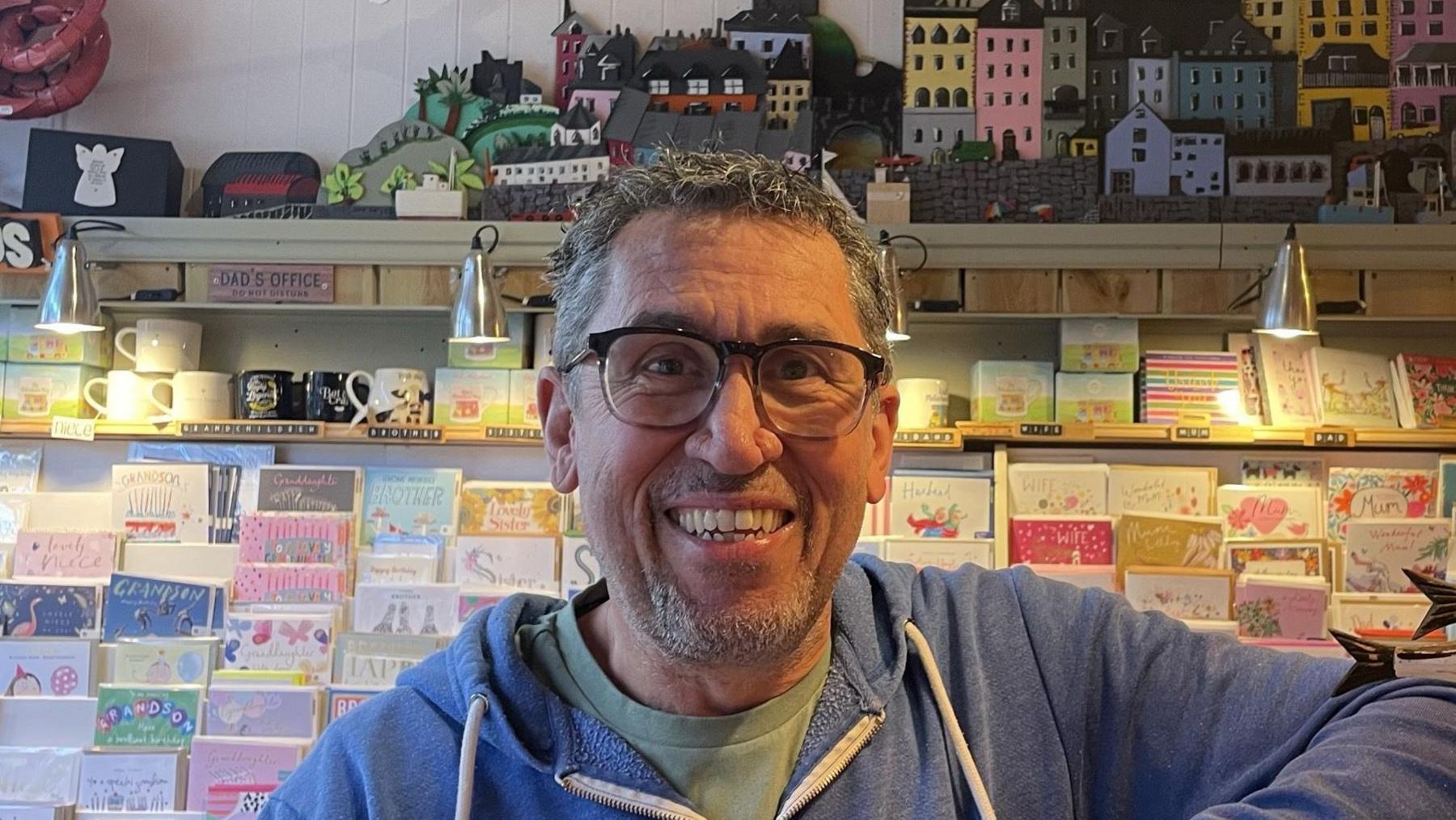 Peter Hall smiles at the camera behind the till of his gift shop. He has short grey hair, is wearing glasses and a blue hoodie. 