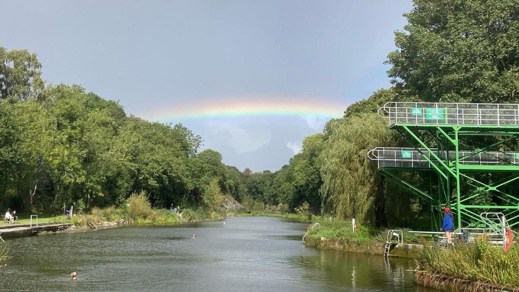 Henleaze Lake is pictured looking down its length from one end, with trees and grassy areas on each side. Some people are swimming in the water and a young woman is standing on the lowest of its three diving boards. There is a rainbow overhead