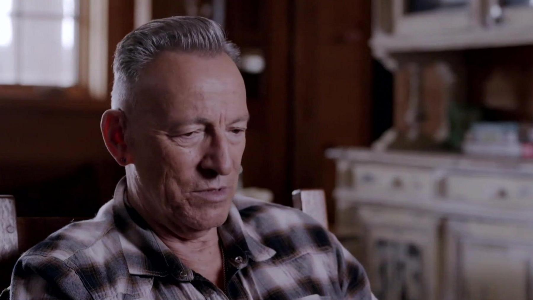 Head and shoulder shot of a middle aged man - rock legend Bruce Springsteen. He has combed-back grey hair, is wearing a purple check shirt, and his expression is serious.