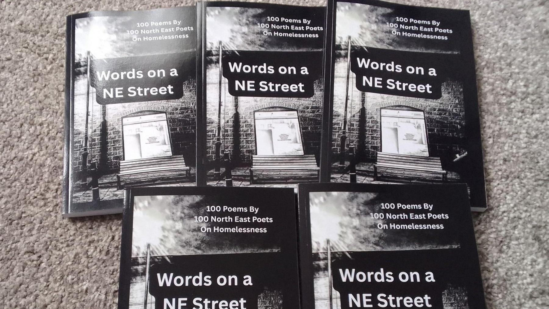 Five of the anthologies are laid out on a grey carpet. The front cover is a black and white picture of a street with a wooden bench on it, along with a lam post and a window to a building where light shines through. The title of the book is: "Words on a NE Street", "100 poems by 100 North East Poets on Homelessness".