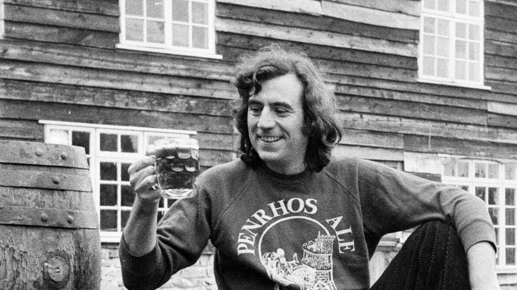 A black and white photo of a man with long dark hair to his shoulders and wearing a top with the word "Penrhos Ale" written on it, around a drawing of a castle. He holds a pint glass filled up liquid up as he looks and smiles at it