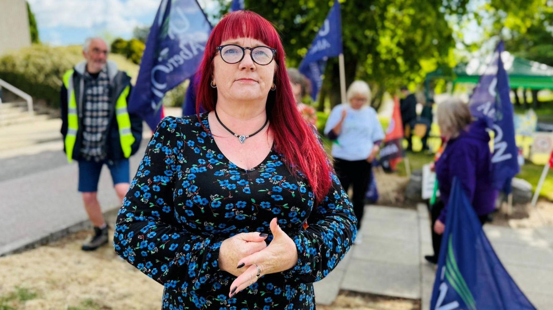 Woman - Inez Kirk from Unison - looking at camera, with bright burgundy hair and a floral blue dress, with people holding flags in the background.