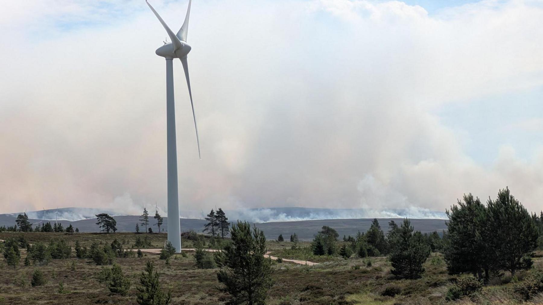 A white wind turbine on an area of moorland with low bushes and pine trees. In the background smoke rises from a wildfire on a hillside.