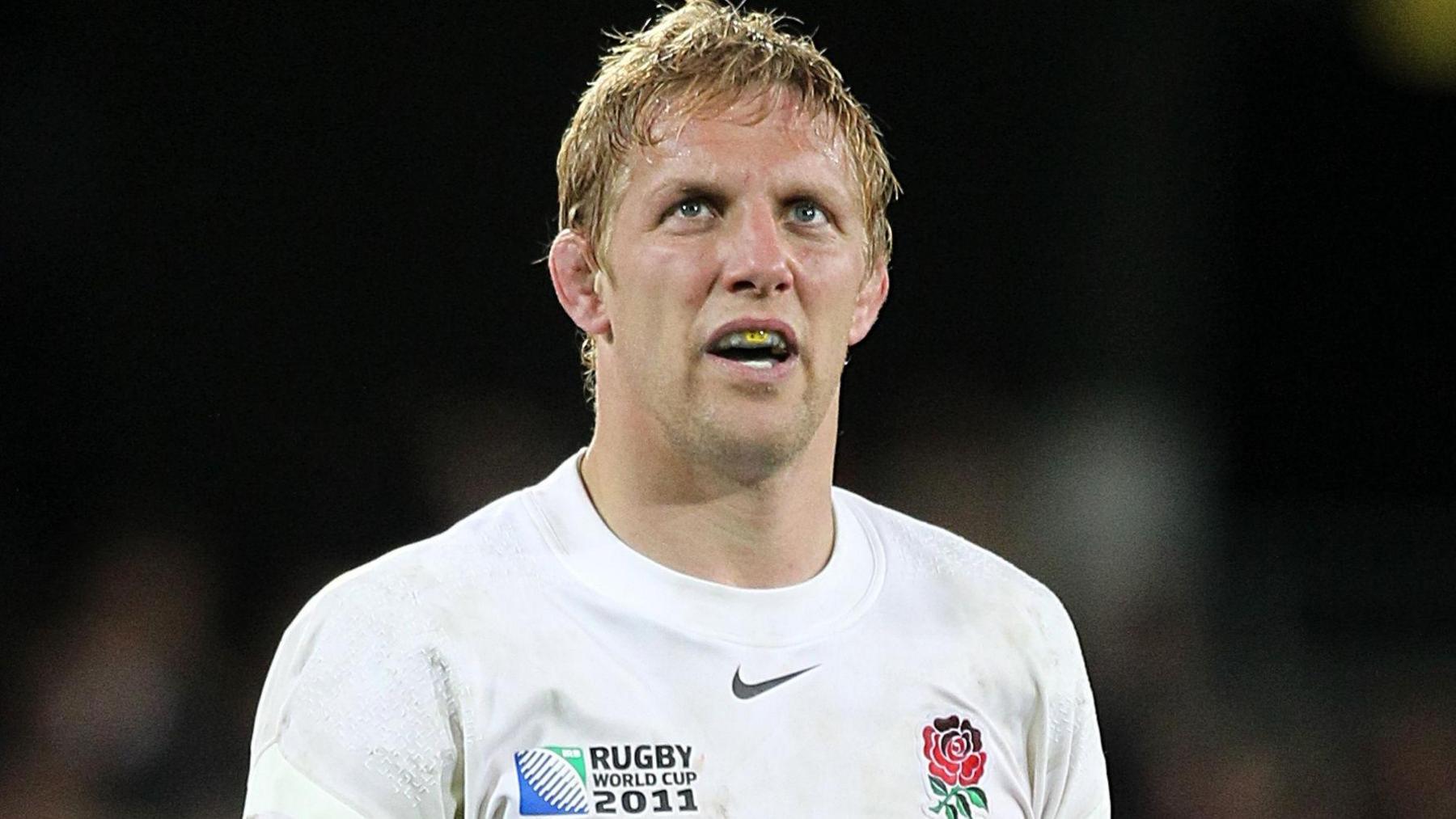 Former England captain and 2003 World Cup winner Lewis Moody pictured on a rugby pitch. He has blonde hair and has a gum shield in his mouth. There is the red rose logo on the right of his white top and 'Rugby World Cup 2011' on the left.
